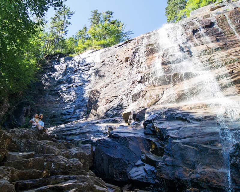 photo of man and child sitting at base of tall rocky waterfall