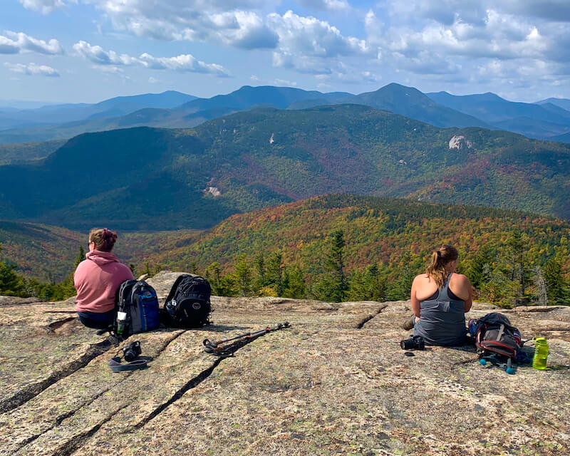 photo of 2 white women sitting on rock looking at mountains